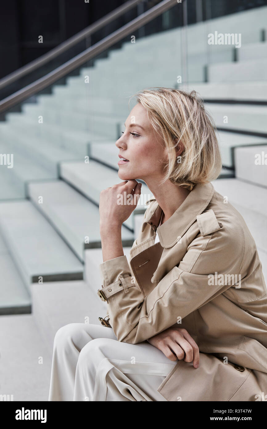 Young blond businesswoman wearing beige trenchcoat sitting on stairs Stock Photo