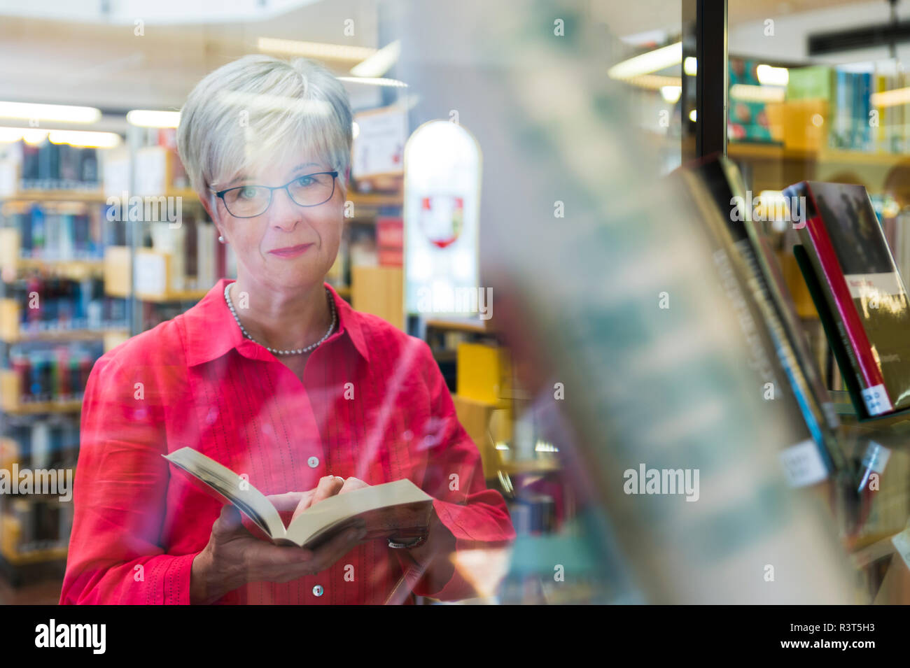 Seniorin mit Buch und Kamerablick hinter einer spiegelnden Glasscheibe.Grevenbroich, Nordrhein-Westfalen, Deutschland Stock Photo