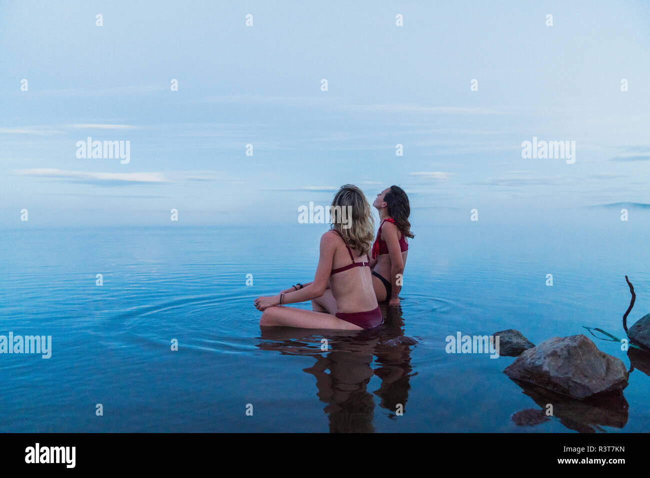 Two young women wearing bikinis, sitting on a stone in a lake Stock Photo