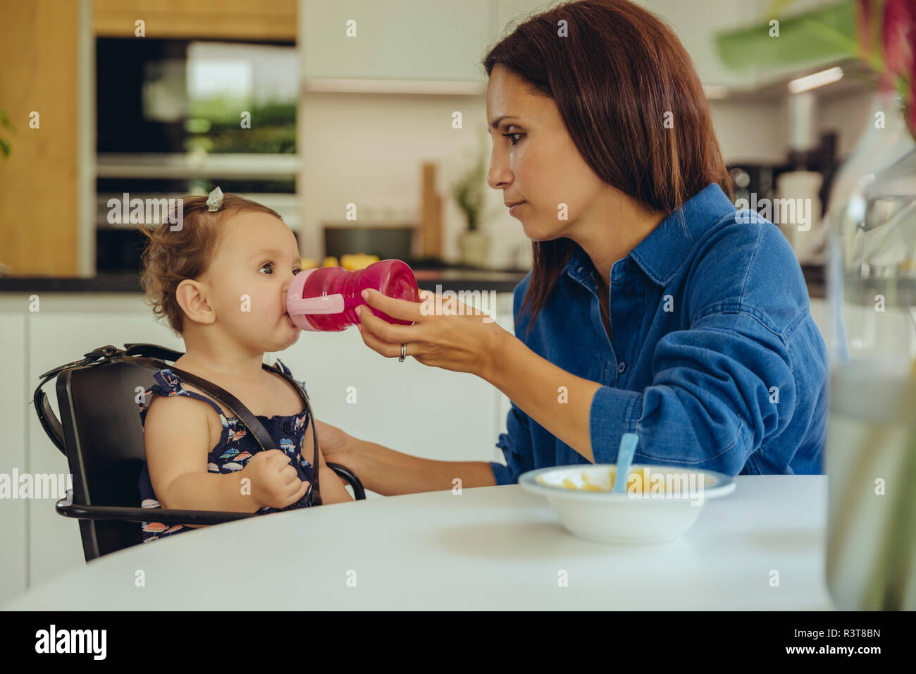 Mother helping baby daughter drinking water from bottle in kitchen Stock Photo