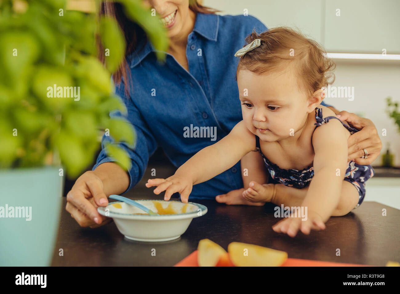Mother with baby daughter eating fruit pulp in kitchen Stock Photo