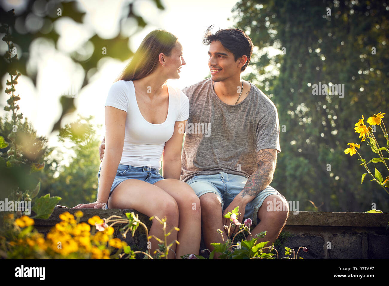 Romantic young couple sitting on wall in a park, with arms around Stock Photo
