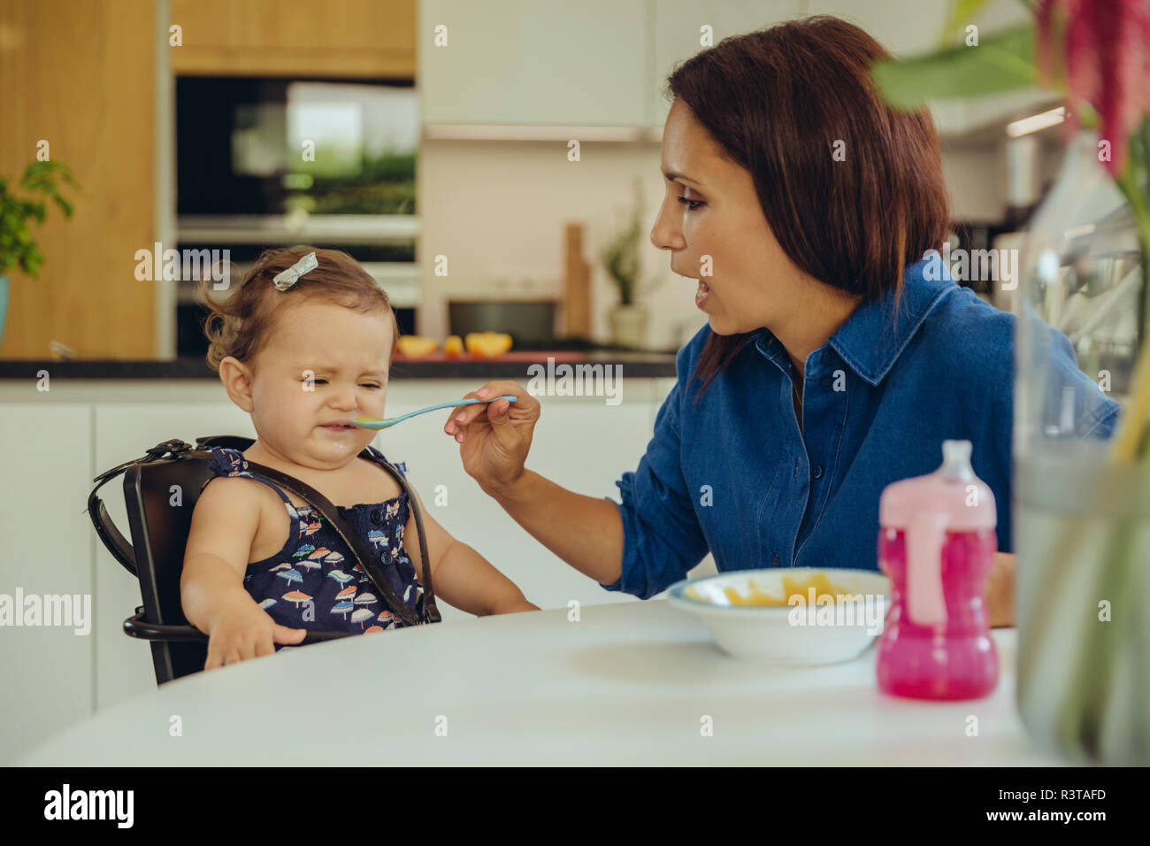 Mother helping disgusted baby daughter eating fruit pulp in kitchen Stock Photo