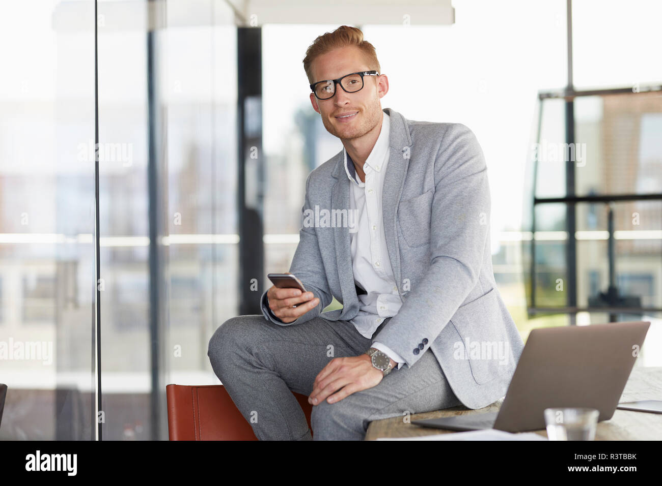 Portrait of smiling businessman with laptop and cell phone sitting on desk in office Stock Photo