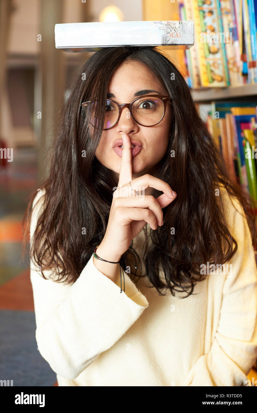 Portrait of young woman with book on top of head asking for silence at the library Stock Photo