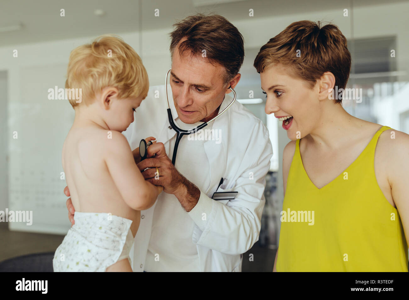 Pediatrician examining toddler with stethoscope, mother standing next to them Stock Photo