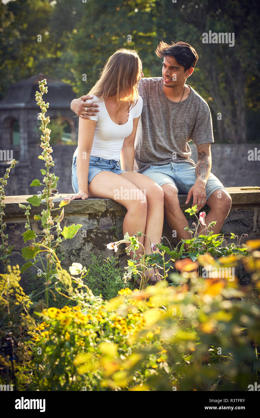 Romantic young couple sitting on wall in a park, with arms around Stock Photo