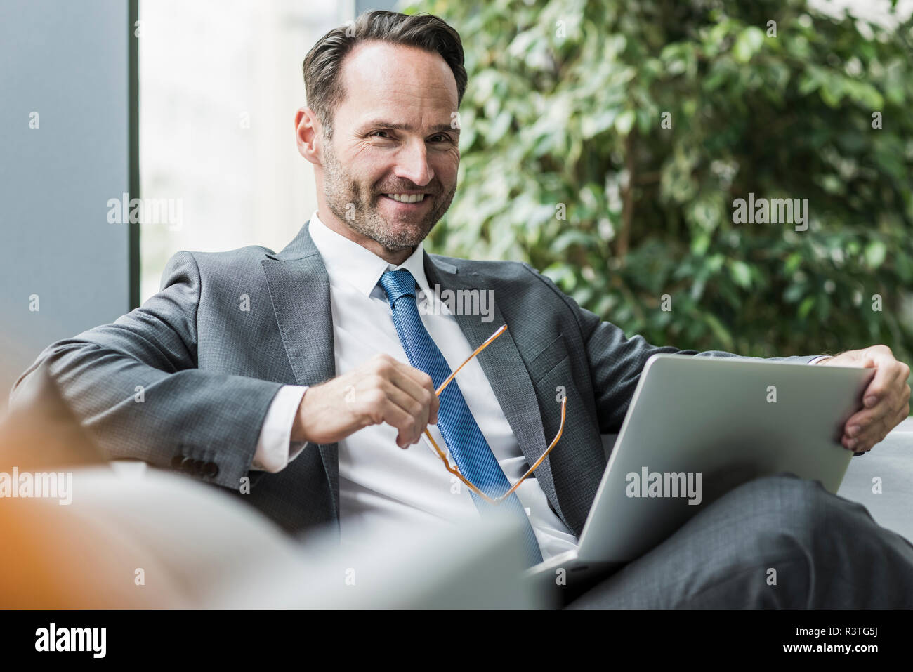 Portrait of smiling businessman sitting in lobby with laptop Stock Photo