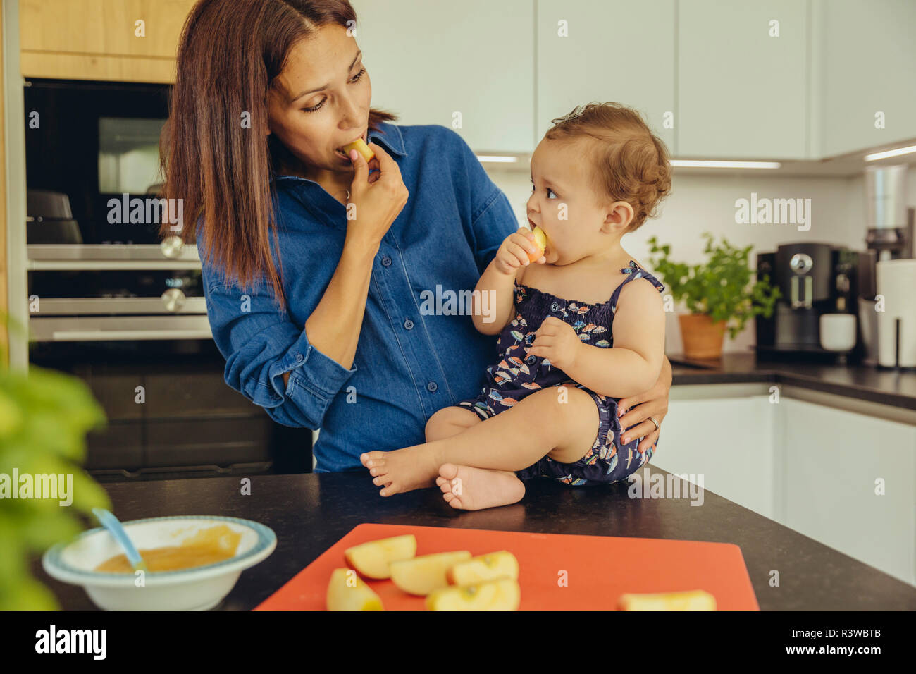 Mother and baby daughter eating apple chunks in kitchen together Stock Photo