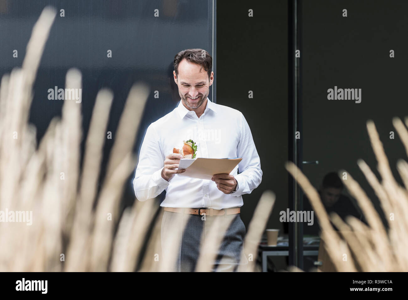 Smiling businessman with sandwich reading documents in front of office building Stock Photo