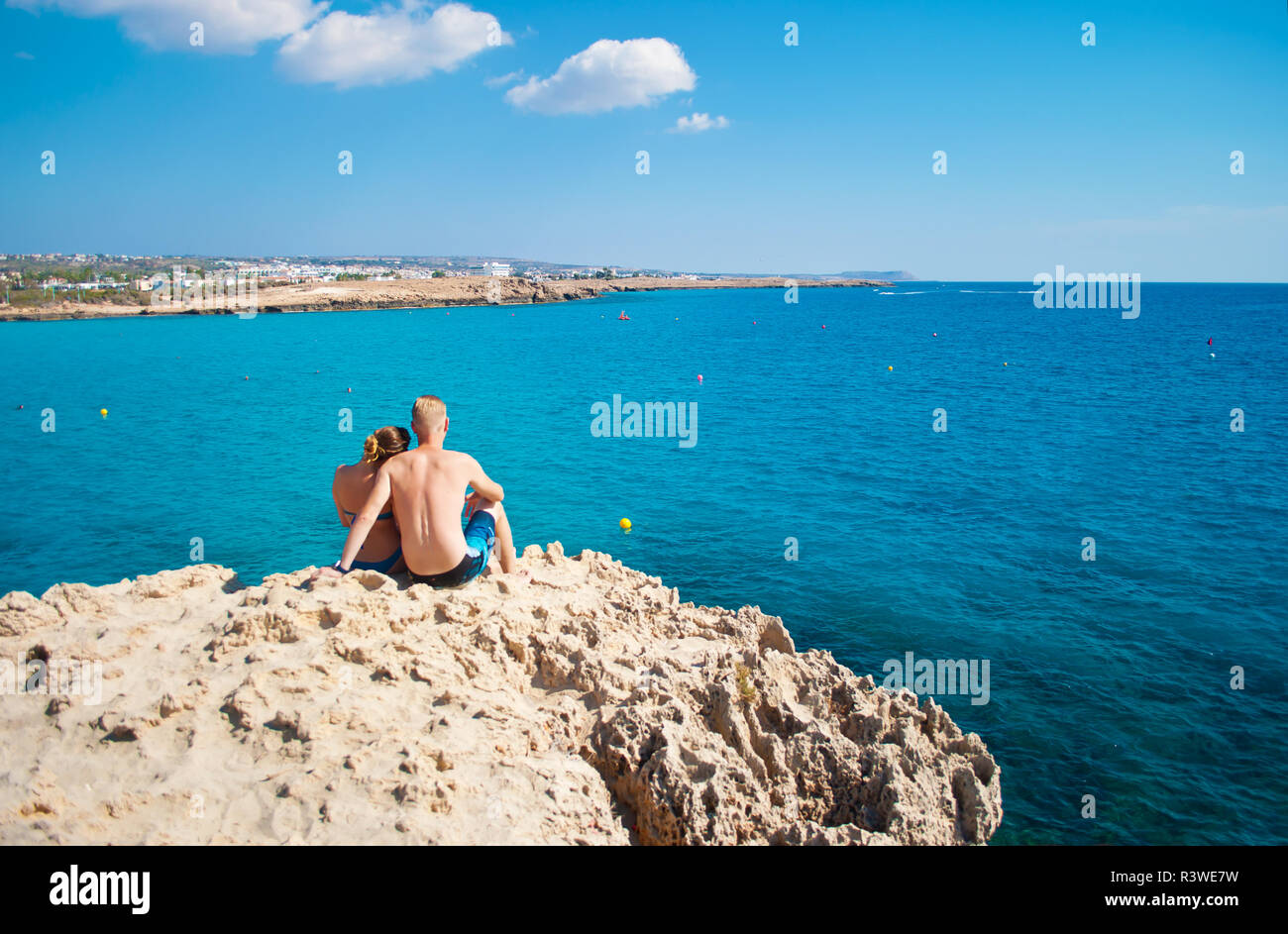 Romantic couple sitting together on a rock near transparent water. Woman leaning her head on man's shoulder. Concept of love, care, tenderness, travel Stock Photo