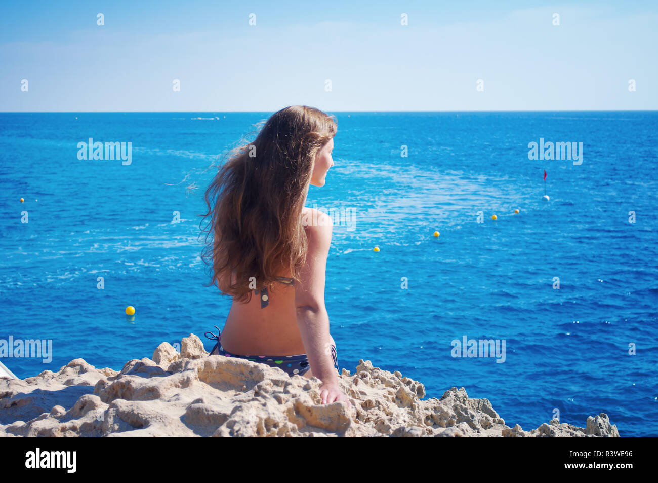 Image of a back of one caucassian girl with long hair sitting on a rock with her hand leaning behind, looking in the distance to the blue sea. Cloudle Stock Photo