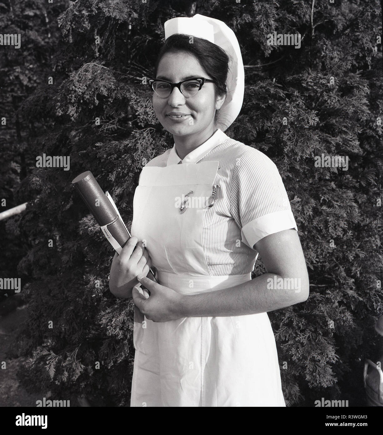 1965, Britain, NHS,a proud overseas nurse from the Indian subcontinent standing outside in her uniform holding her British Royal College of Nursiing (RCN) qualifications and wearing a fob watch and nursing qualification badge, England, UK. Stock Photo