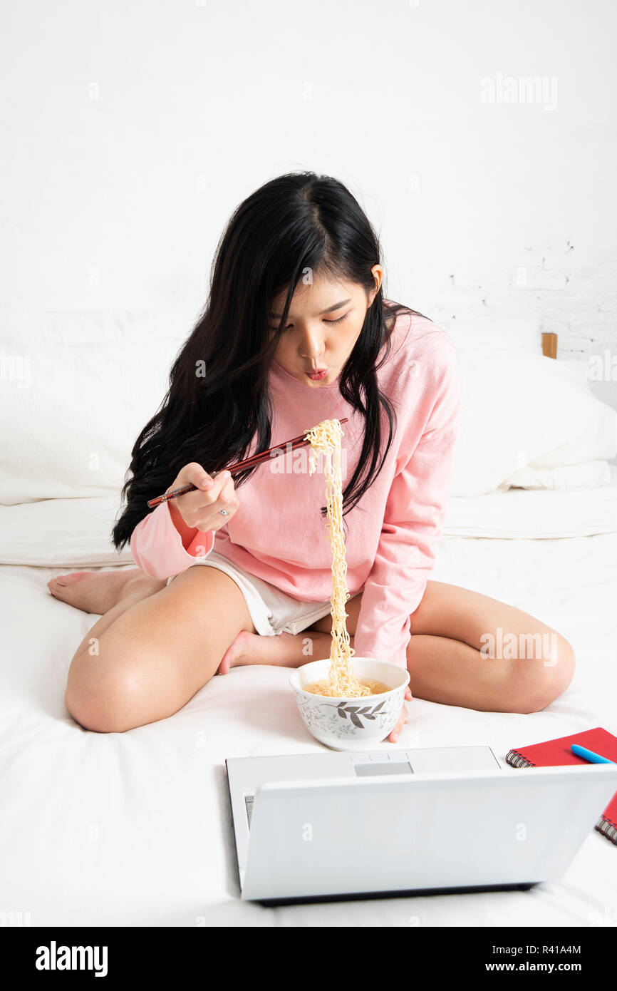 Girl eating instant noodle in hurry time while running her business at home on notebook computer in bedroom. Stock Photo