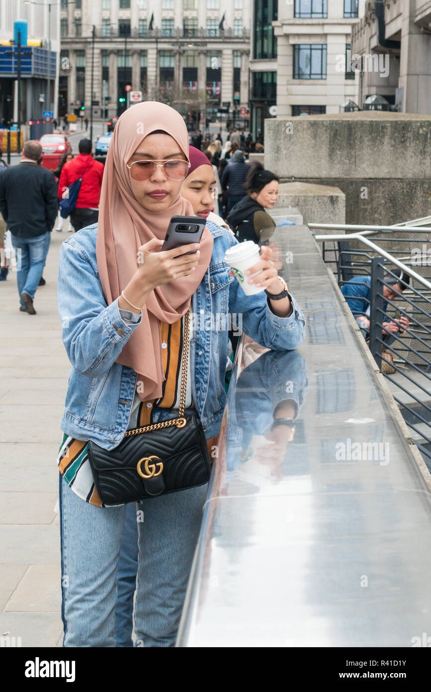 denim jeans and jacket, dull spring day, gg handbag purse, headscarf, London uk, muslim woman taking a photograph Stock Photo