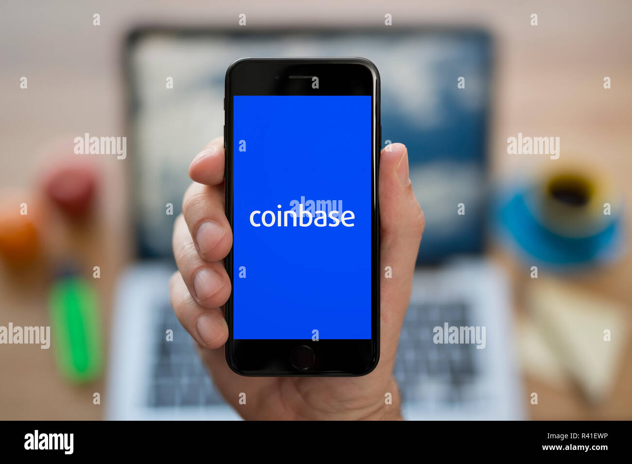 A man looks at his iPhone which displays the Coinbase logo, while sat at his computer desk (Editorial use only). Stock Photo