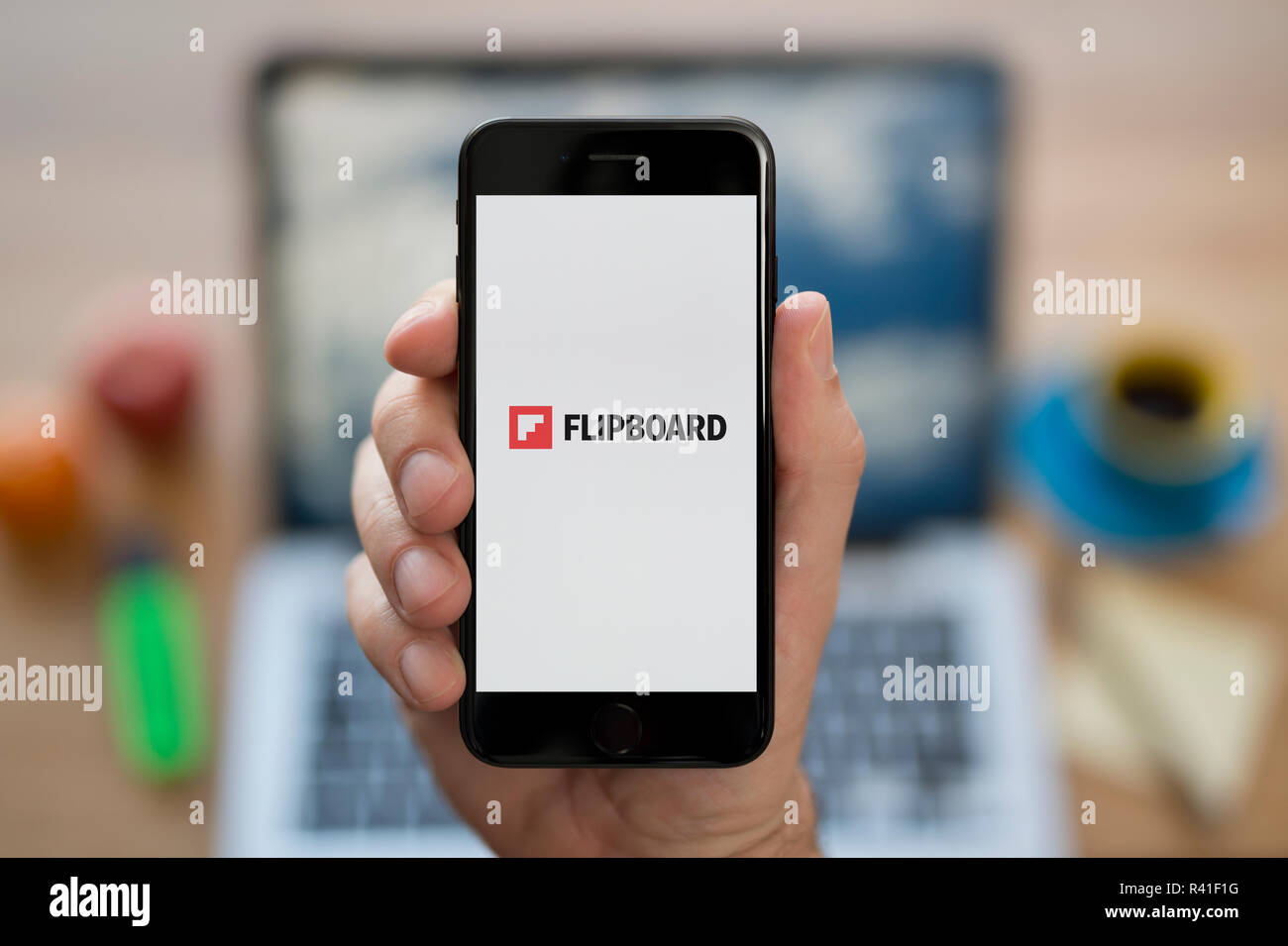 A man looks at his iPhone which displays the Flipboard logo, while sat at his computer desk (Editorial use only). Stock Photo