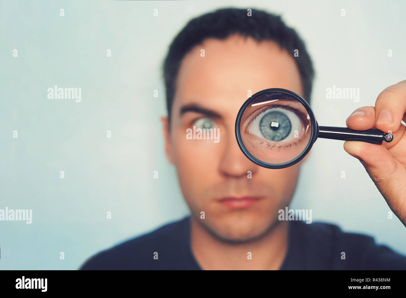 Potrait of young man looking through magnifying glass on white blurred background. View to male blue eye through the lens. Loupe with macro eye in person's hand on the foreground. Information search. Stock Photo