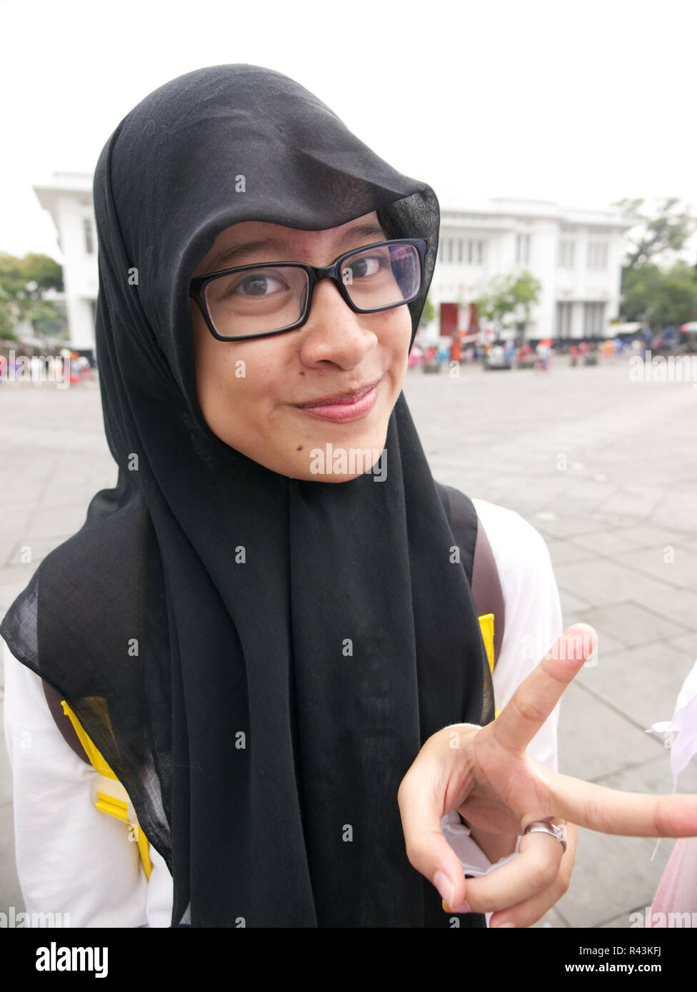 Vertical portrait of Indonesian teenager wearing black headscarf and white dress and glasses with grin making peace sign in Jakarta square. Stock Photo