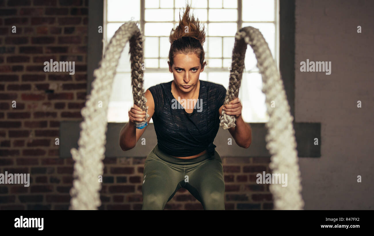 Athlete moving the ropes in wave motion as part of fat burning workout in fitness studio. Sportswoman exercising with battling ropes at gym. Stock Photo