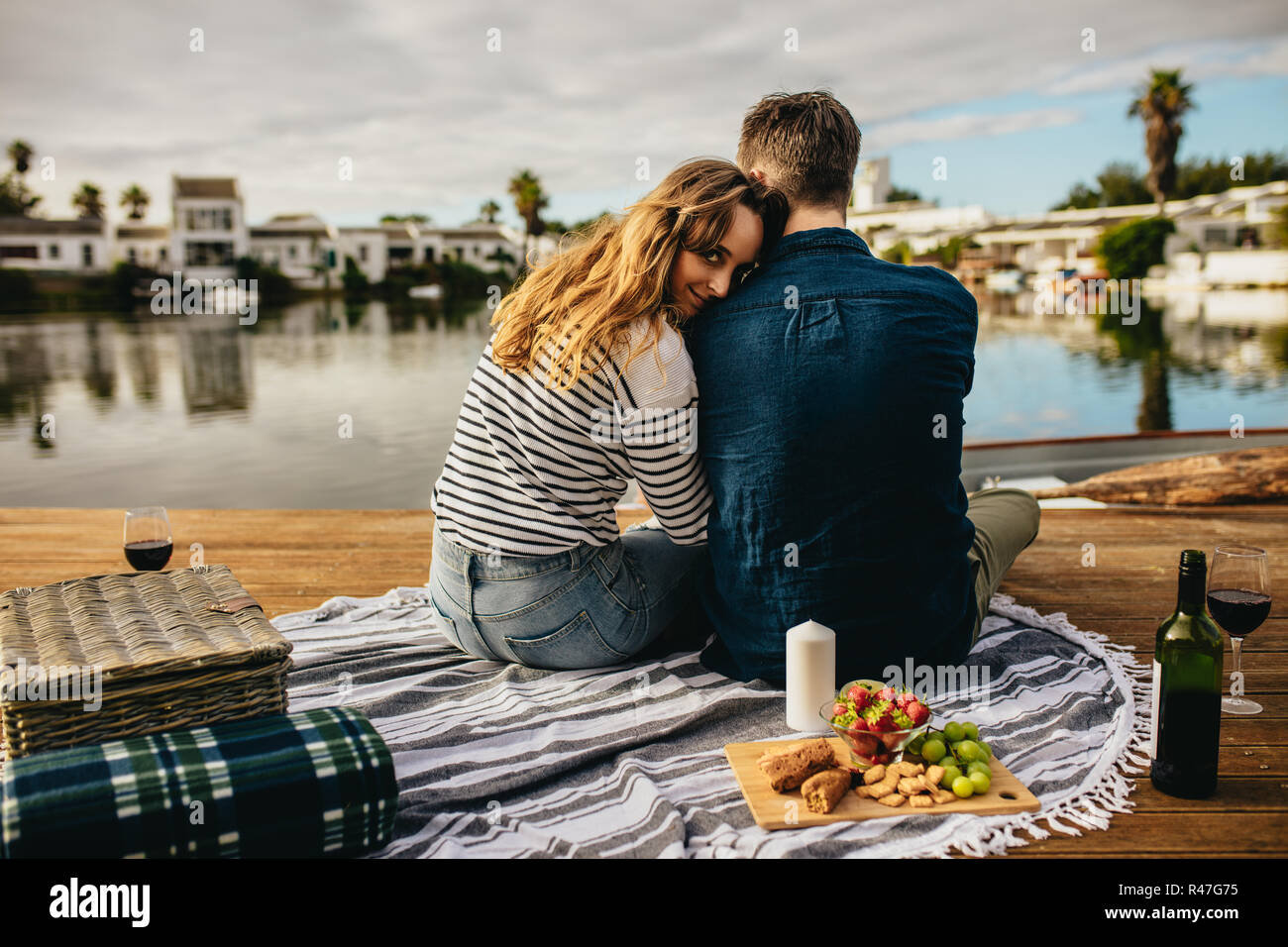 Rear view of a couple in love sitting side by side beside a lake. Couple on a day out sitting together with picnic food and drinks facing the lake wit Stock Photo