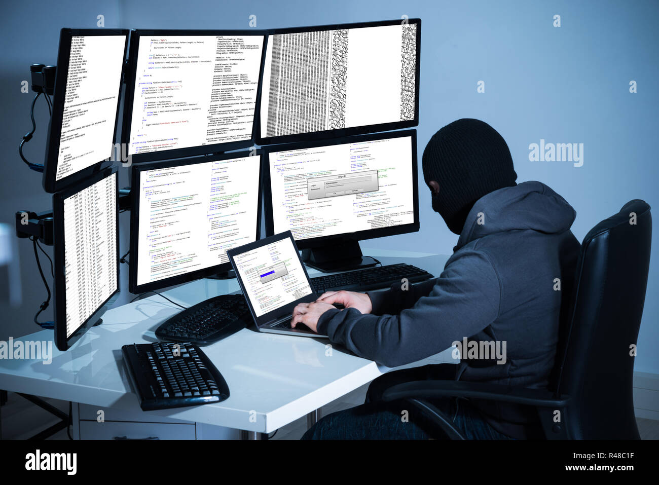 Hacker Using Laptop Against Multiple Monitors At Desk Stock Photo