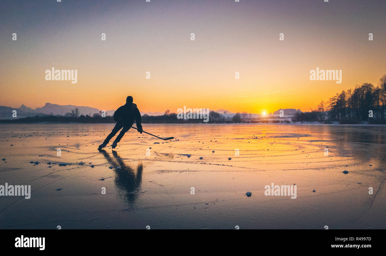 Scenic panoramic view of the silhouette of a young hockey player skating on a frozen lake with amazing reflections in beautiful golden evening light Stock Photo