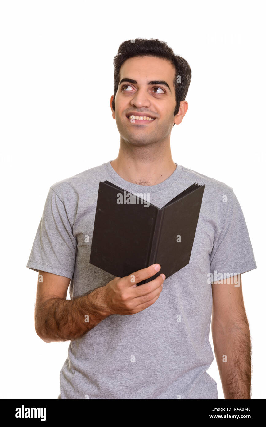 Young happy Persian man holding book while thinking Stock Photo