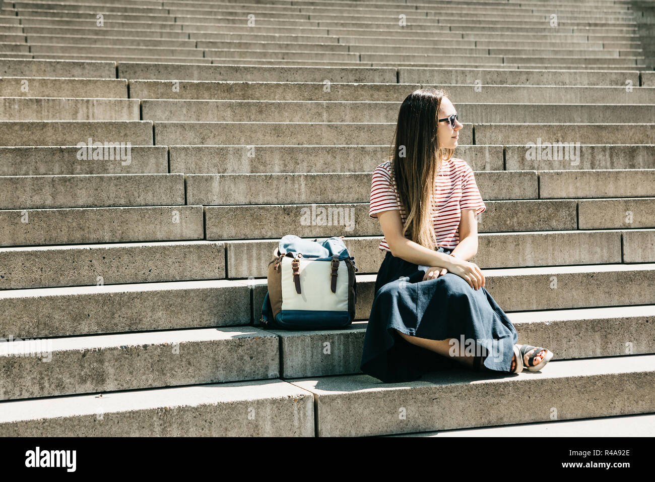 Beautiful tourist girl or student in sunglasses and with a backpack sits on the stairs and rests. Lifestyle Stock Photo