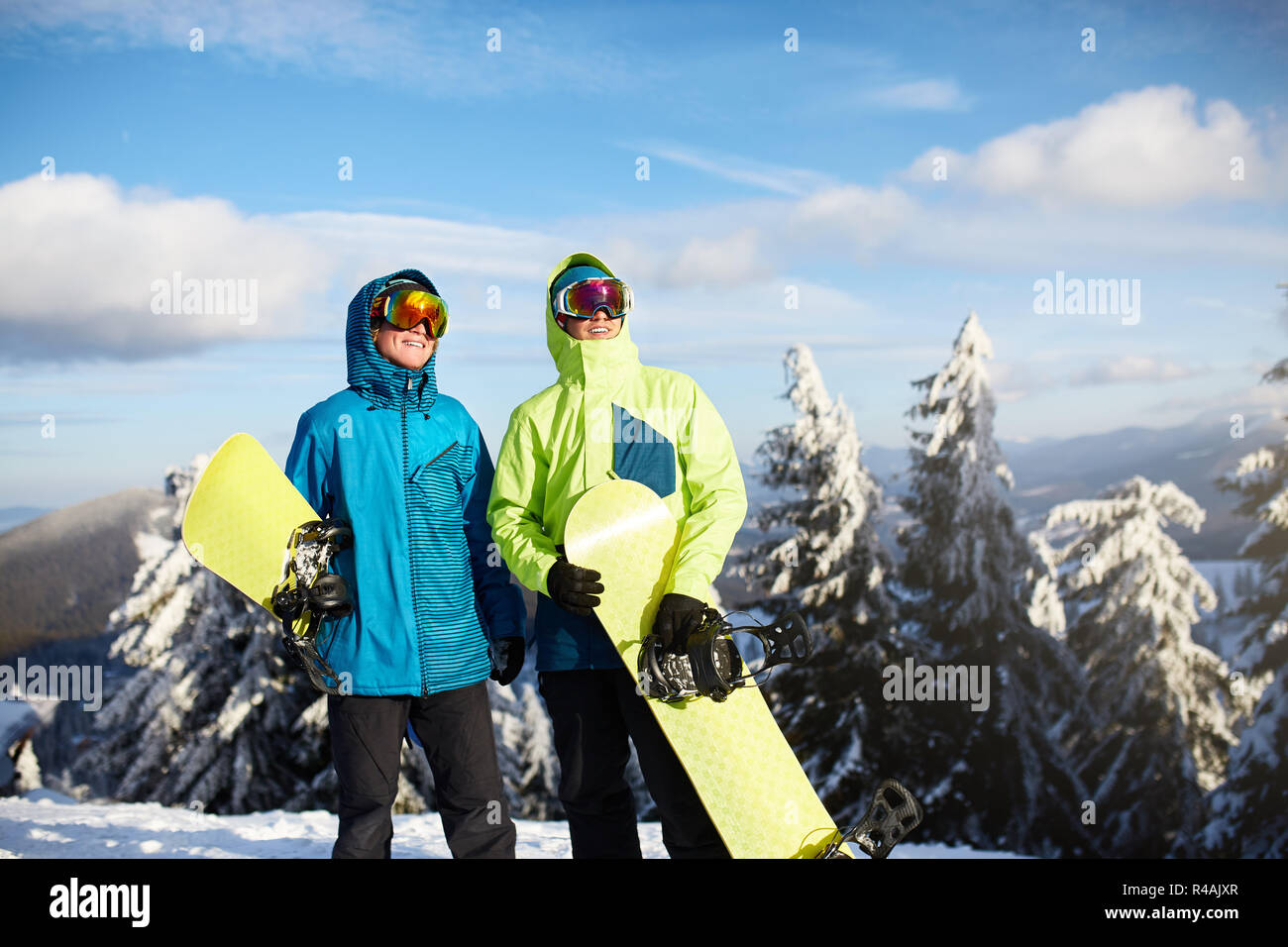 Two snowboarders posing at ski resort. Riders friends carrying their snowboards through forest for backcountry freeride and wearing reflective goggles, colorful fashion outfit. Copyspace area. Stock Photo