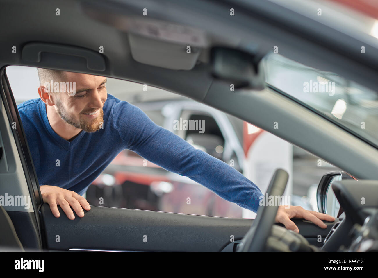 Handsome man in blue sweater looking at indoor design of luxury car. Brunette young customer coming to car salon and wanting buying vehicle. Happy driver smiling and leaning on front auto door. Stock Photo