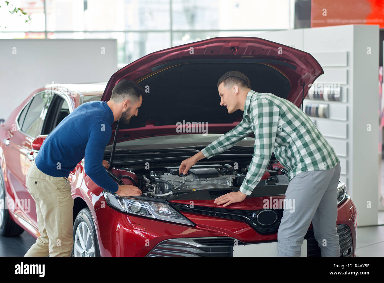 Two attractive men standing back to camera under car hood. Brothers in casual clothes talking near red auto and fixing it. Young man coming to salon helping his friend choosing new vehicle. Stock Photo