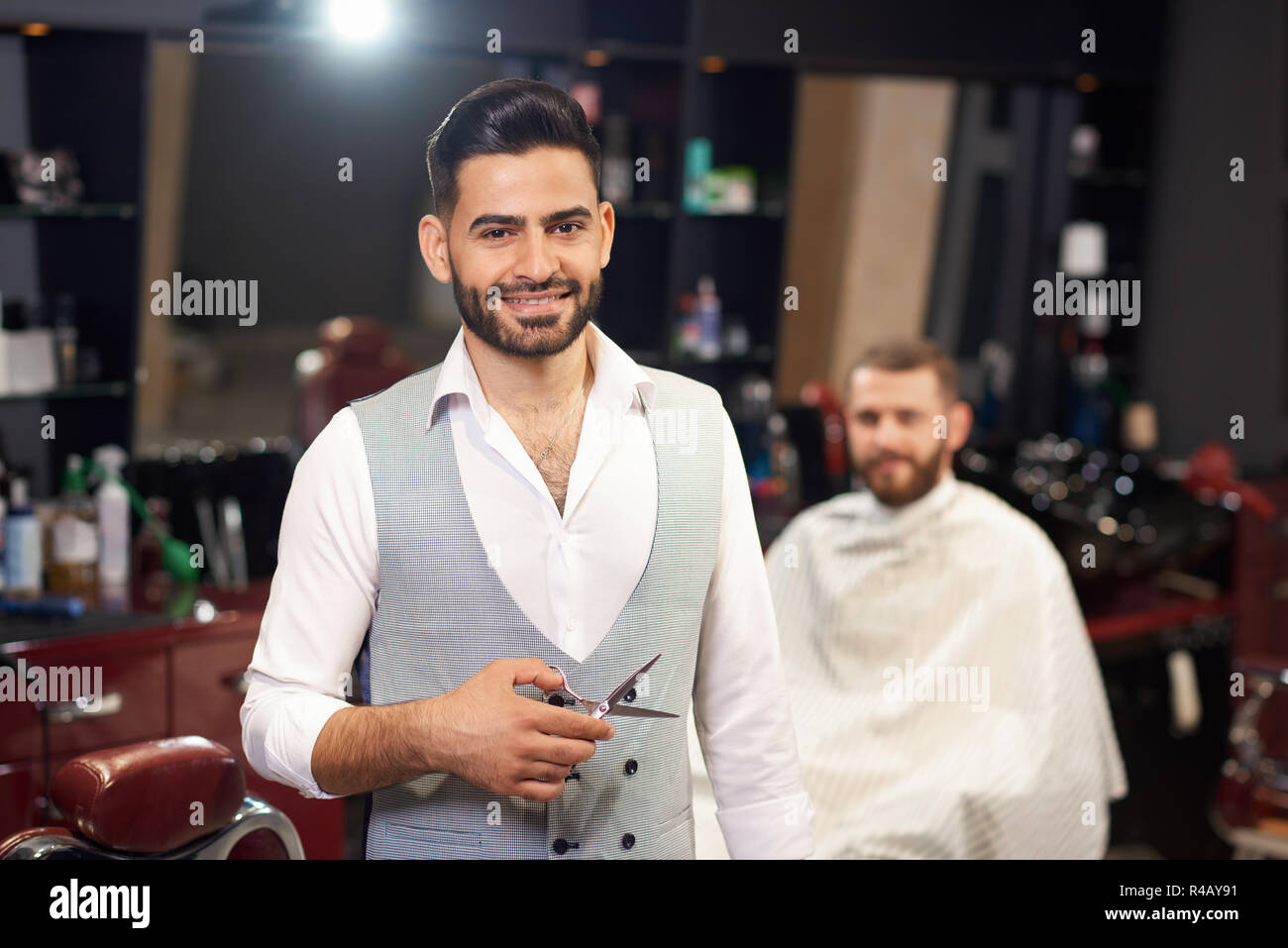 Portrait of professional barber looking at camera, smiling and posing. Stylish man keeping scissors during doing haircut in barber shop. Smiling male customer sitting in chair at background. Stock Photo