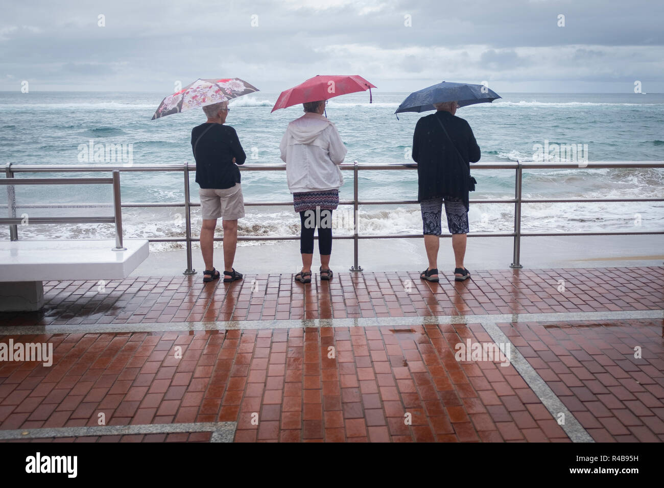 Three people with their umbrellas observe the landscape in the rain in the beach of Las Canteras in Las Palmas de Gran Canaria, Spain Stock Photo