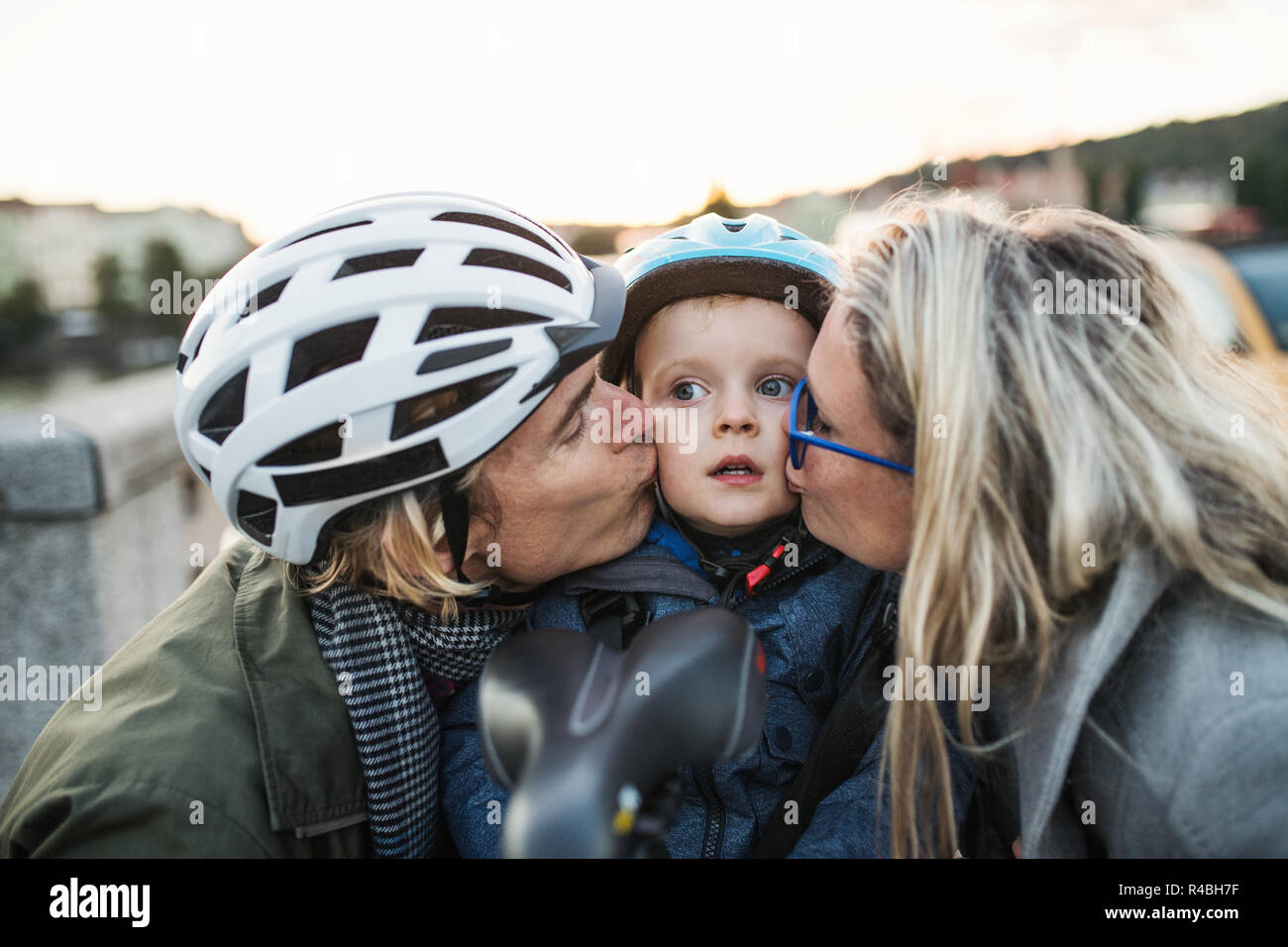 A small toddler boy with helmet and young parents outdoors in city, kissing. Stock Photo