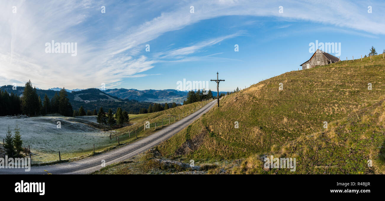 open country landscape with grassy meadow and road and rime and ice and old wooden cross in the middle under an expansive sky and a wooden hut up on t Stock Photo