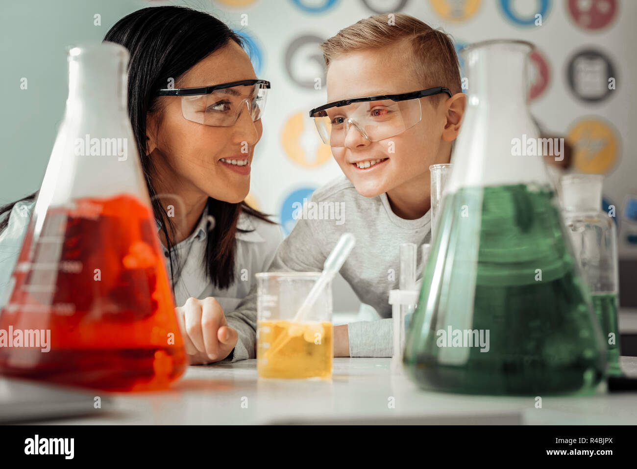 Practical lesson. Kind blonde boy keeping smile on his face while looking at test tubes with reagents Stock Photo