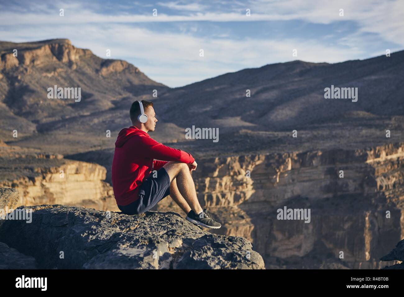 Relaxation in mountains. Young man with headphones sitting on the edge of cliff and listening music. Jebel Akhdar, Grand Canyon of Oman. Stock Photo