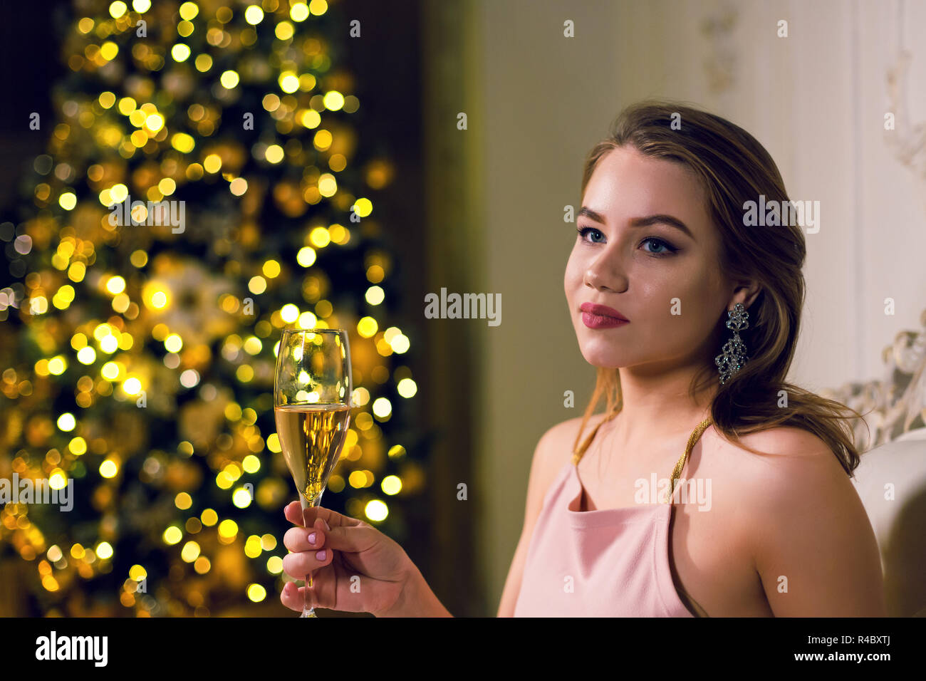 Portrait of a beautiful young woman is sitting in a dark chic hall with a Christmas tree dressed up in a garland in ampir chair with a glass of champa Stock Photo