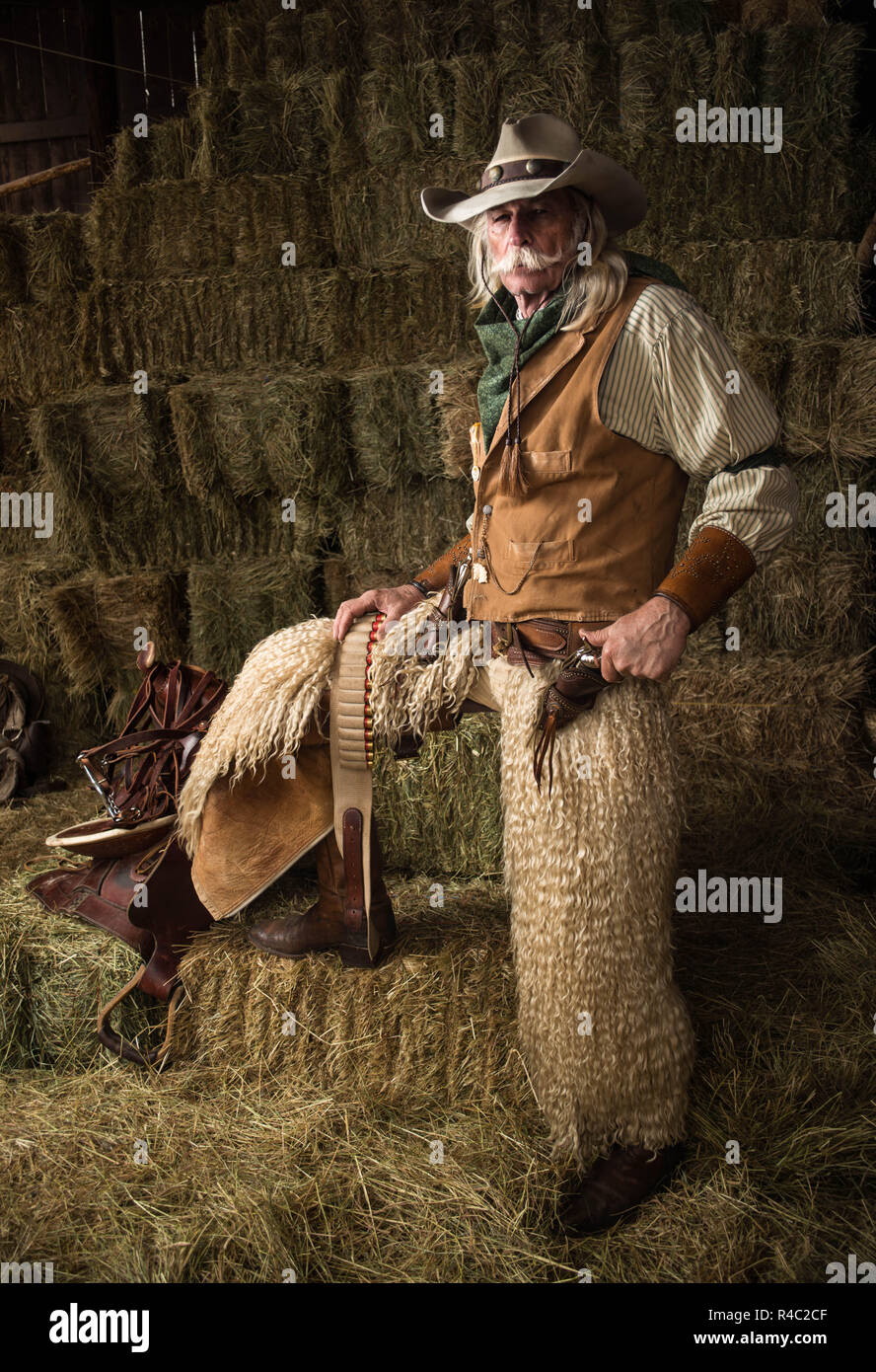 Authentic old west cowboy portrait with wooly chaps, pistol, cowboy hat, leather vest, bandanna in stable Stock Photo