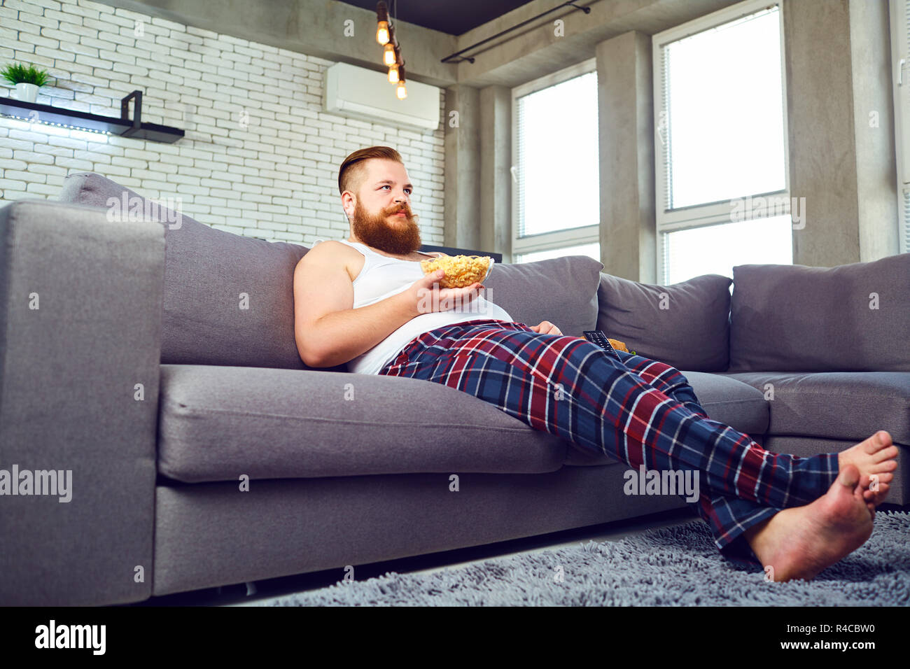 Thick bearded man eating popcor sitting on the couch. Stock Photo