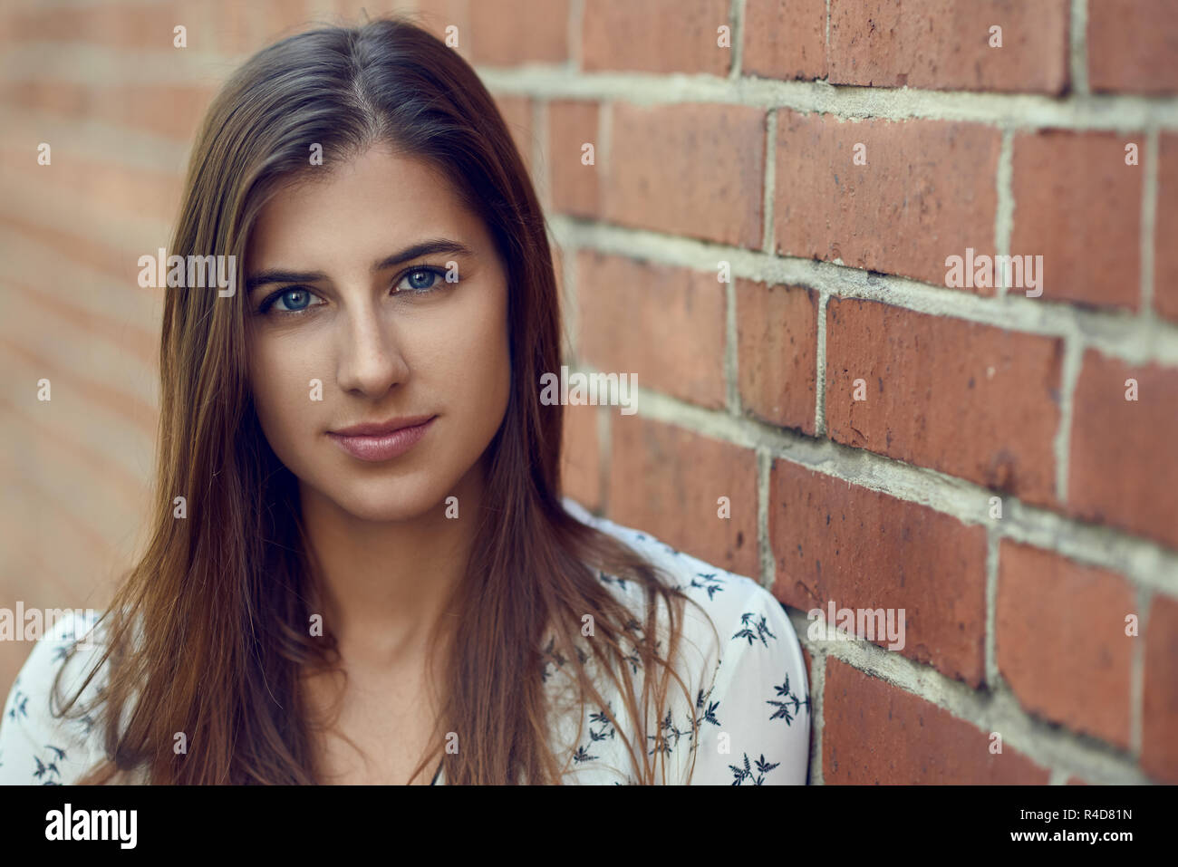 Portrait of young beautiful brunette woman with friendly pretty face, in white blouse, looking at camera while standing near red brick wall with copy  Stock Photo