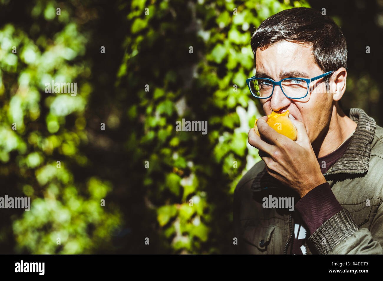 Young man with glasses and blue eyes biting a fruit, peach, with green leaves background Stock Photo