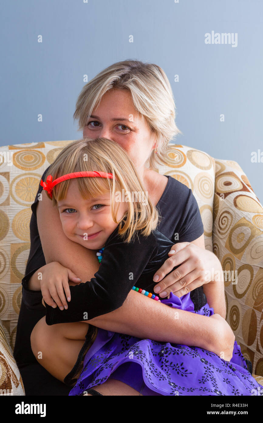 Loving mother hugging her cute little daughter as she sits smiling at the camera while relaxing in a comfortable armchair at home Stock Photo