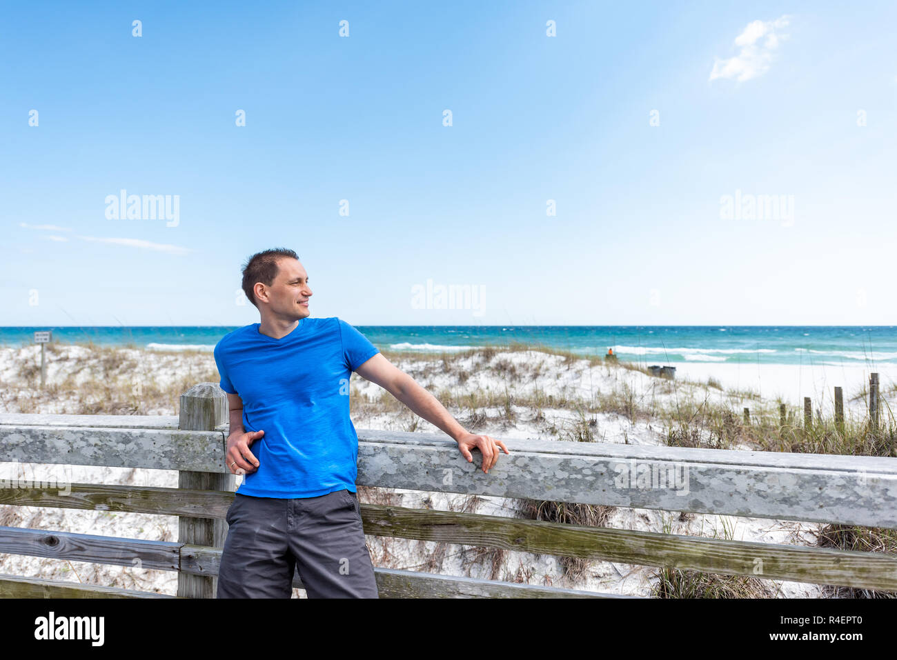 Destin, USA Miramar beach city town village day in Florida panhandle gulf of mexico ocean water, young happy man in blue shirt leaning on wooden fence Stock Photo