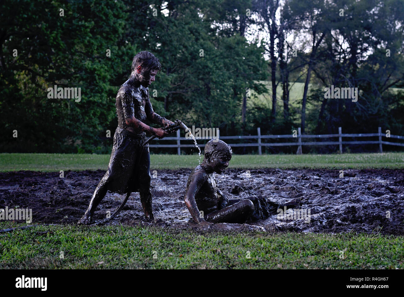 one muddy boy cleaning off another with water from the garden hose after a mud fight Stock Photo
