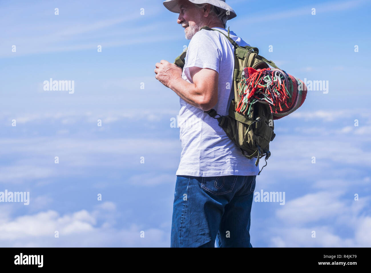 Old man traveler portrait enjoy the clouds view on the top - backpack and colored retro style blanket on the back. Happy lifestyle and modern people - Stock Photo