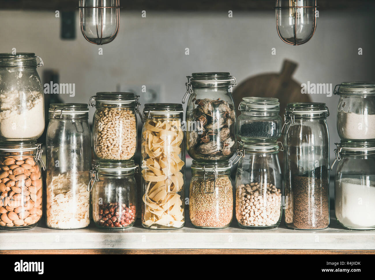 Rustic kitchen food storage arrangement. Various grains, cereals, nuts, dry fruit, flour and pasta kinds in glass jars over concrete kitchen counter. Stock Photo