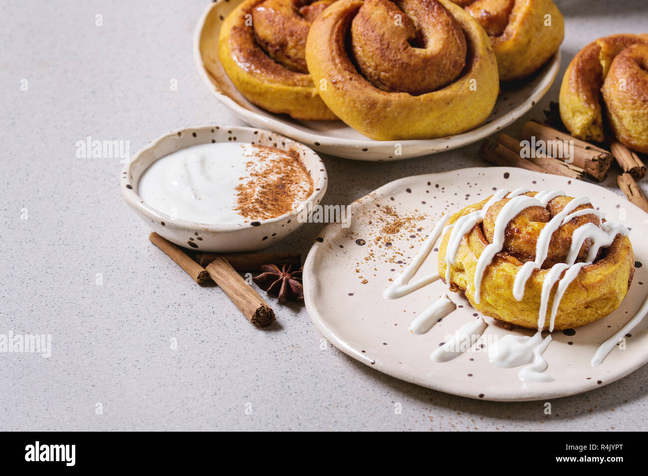 Homemade pumpkin cinnamon bun rolls sweet autumn baked dessert with cream cheese sauce in spotted ceramic plates with cinnamon sticks over white marbl Stock Photo