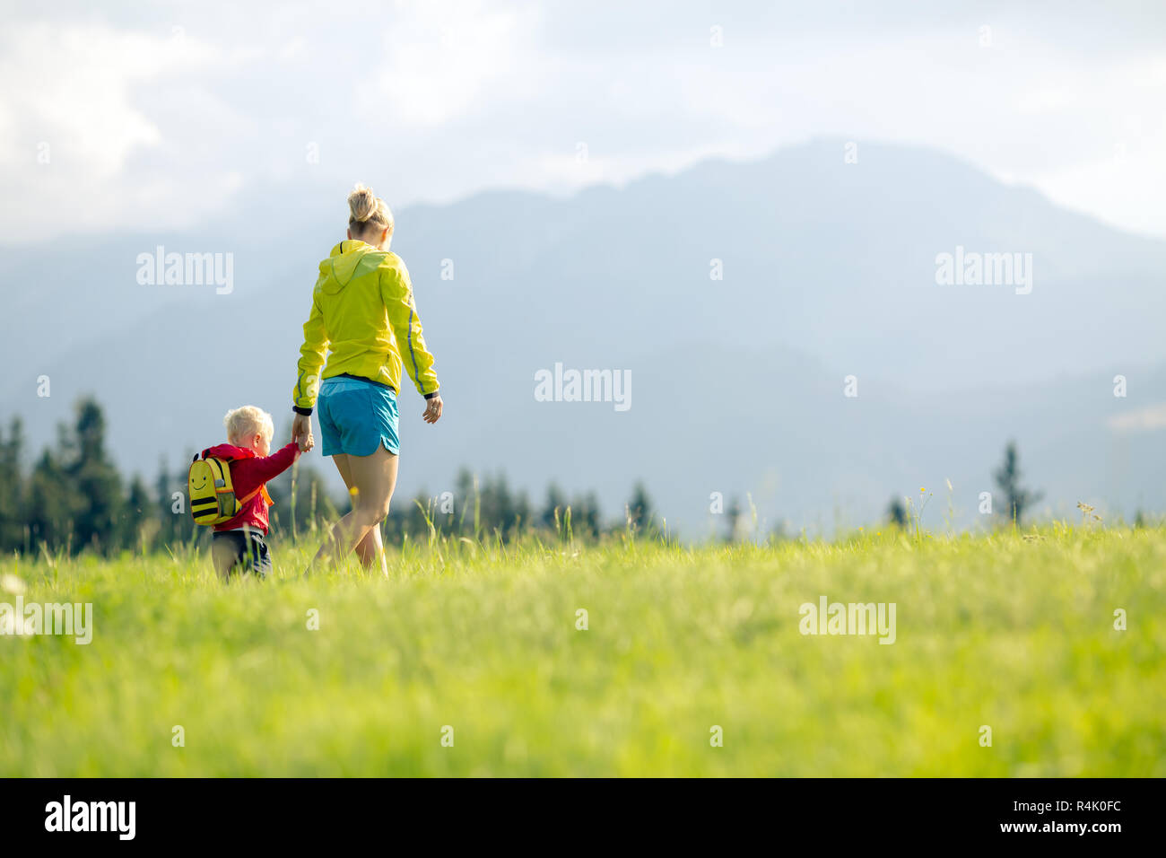 Happy mother with baby boy walking on green meadow. Hiking adventure with child on summer family trip in mountains. Vacations or weekend activity with Stock Photo